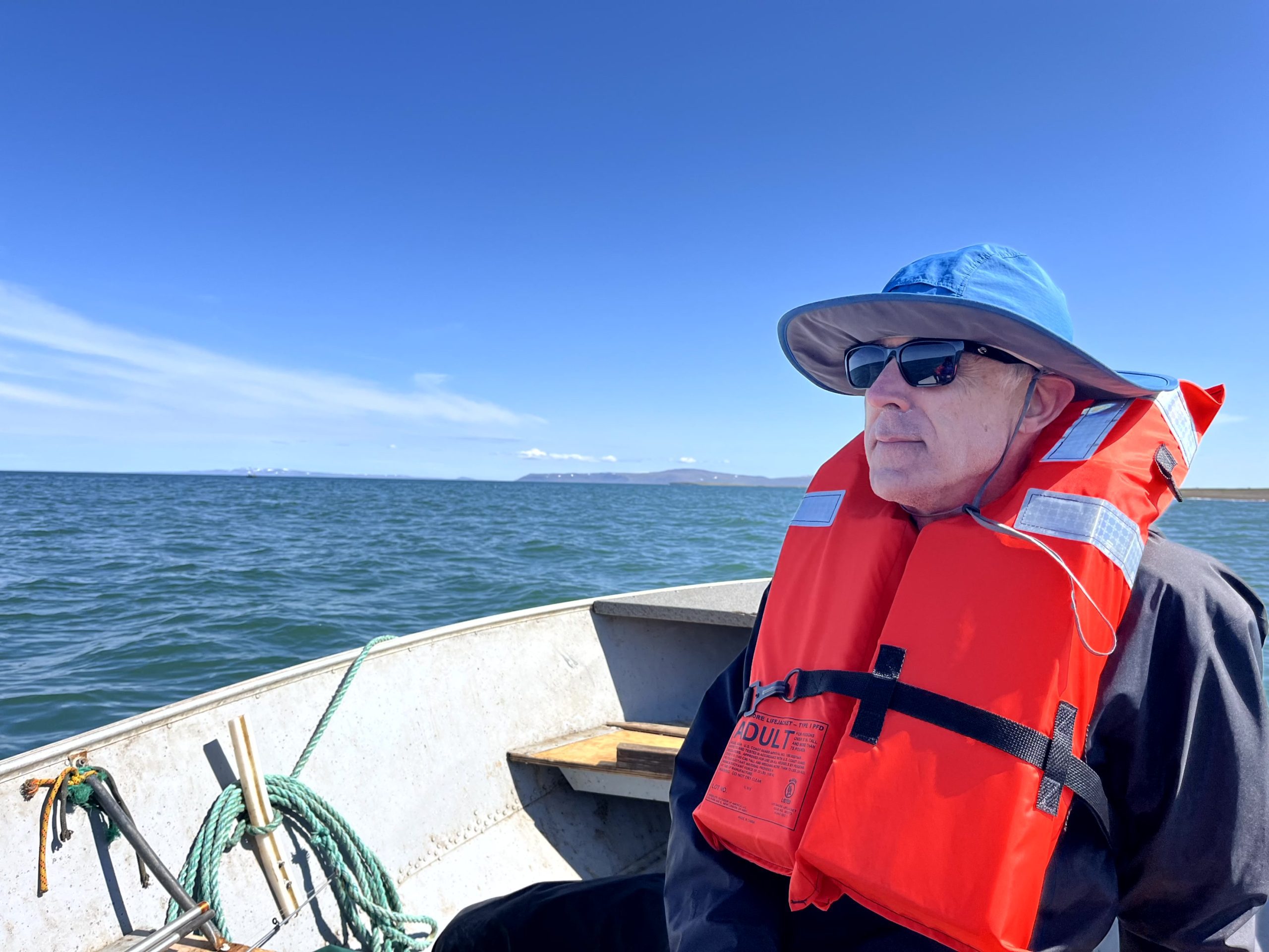 Someone sits in a boat wearing a life vest. The water around them is calm and blue. The sky is blue and partially cloudy. A sliver of land can be seen along the horizon.