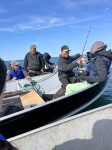People sit in boats lined up with each other. One of them is holding a fishing rod and lifting up a fish. The water around them is calm and blue. The sky is blue with wispy clouds.