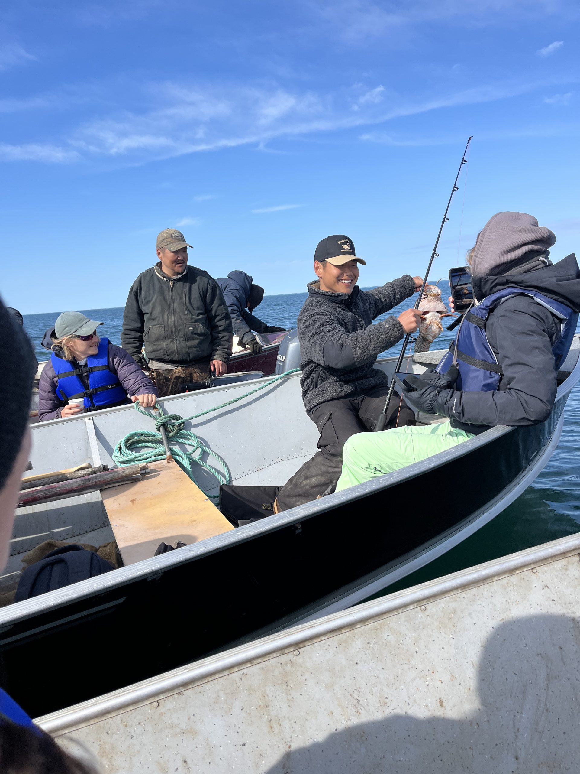 People sit in boats lined up with each other. One of them is holding a fishing rod and lifting up a fish. The water around them is calm and blue. The sky is blue with wispy clouds.