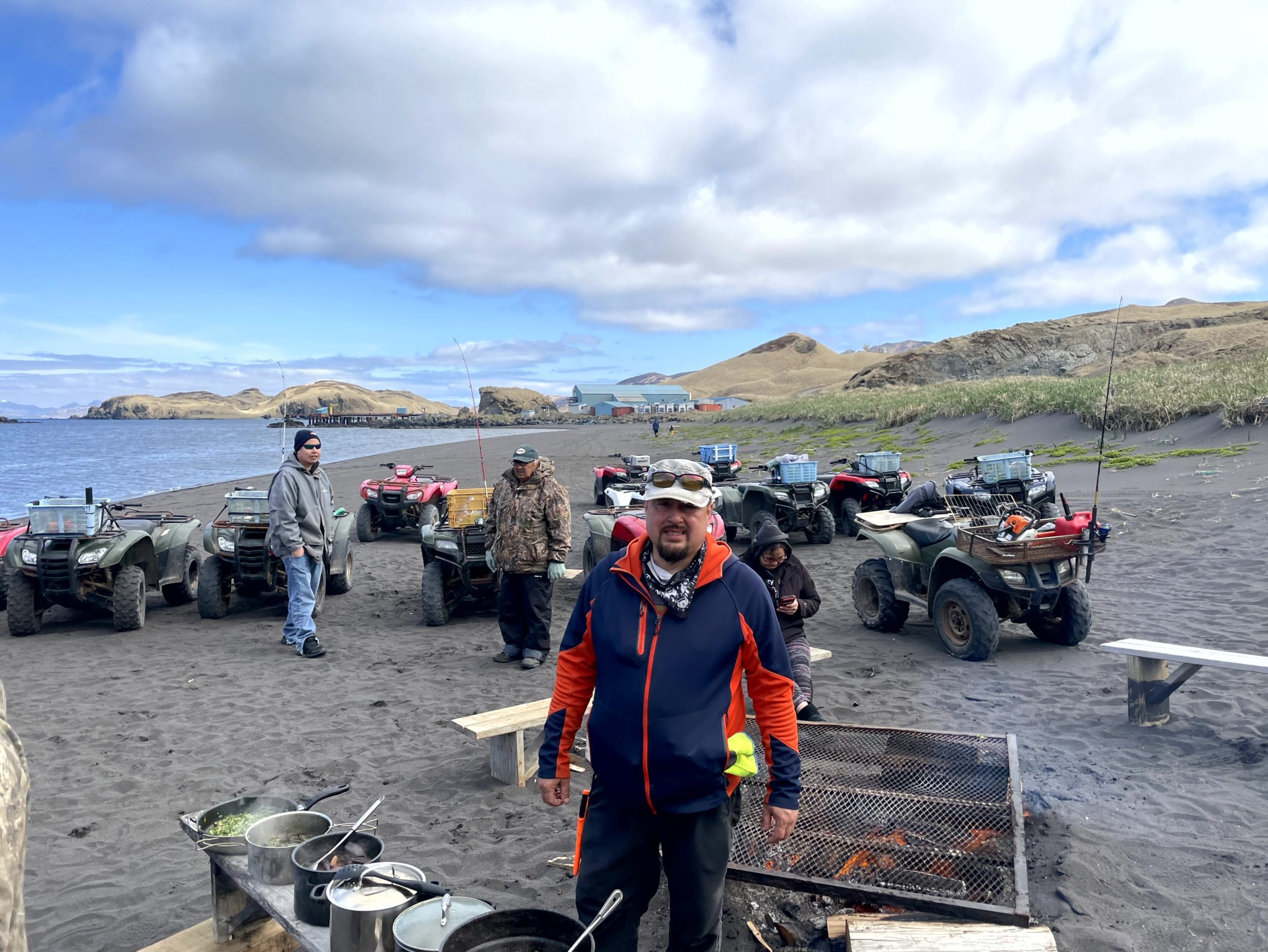 Alex stands next to a fire pit, behind him are a bunch of ATV's and three people. They are on a beach, there are grassy hills in the background and the sky is partially cloudy