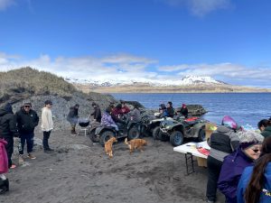 A group of people stand among ATV's and a grill. Behind them are snow-capped mountains and a calm, blue ocean. The sky is partially cloudy and very blue.