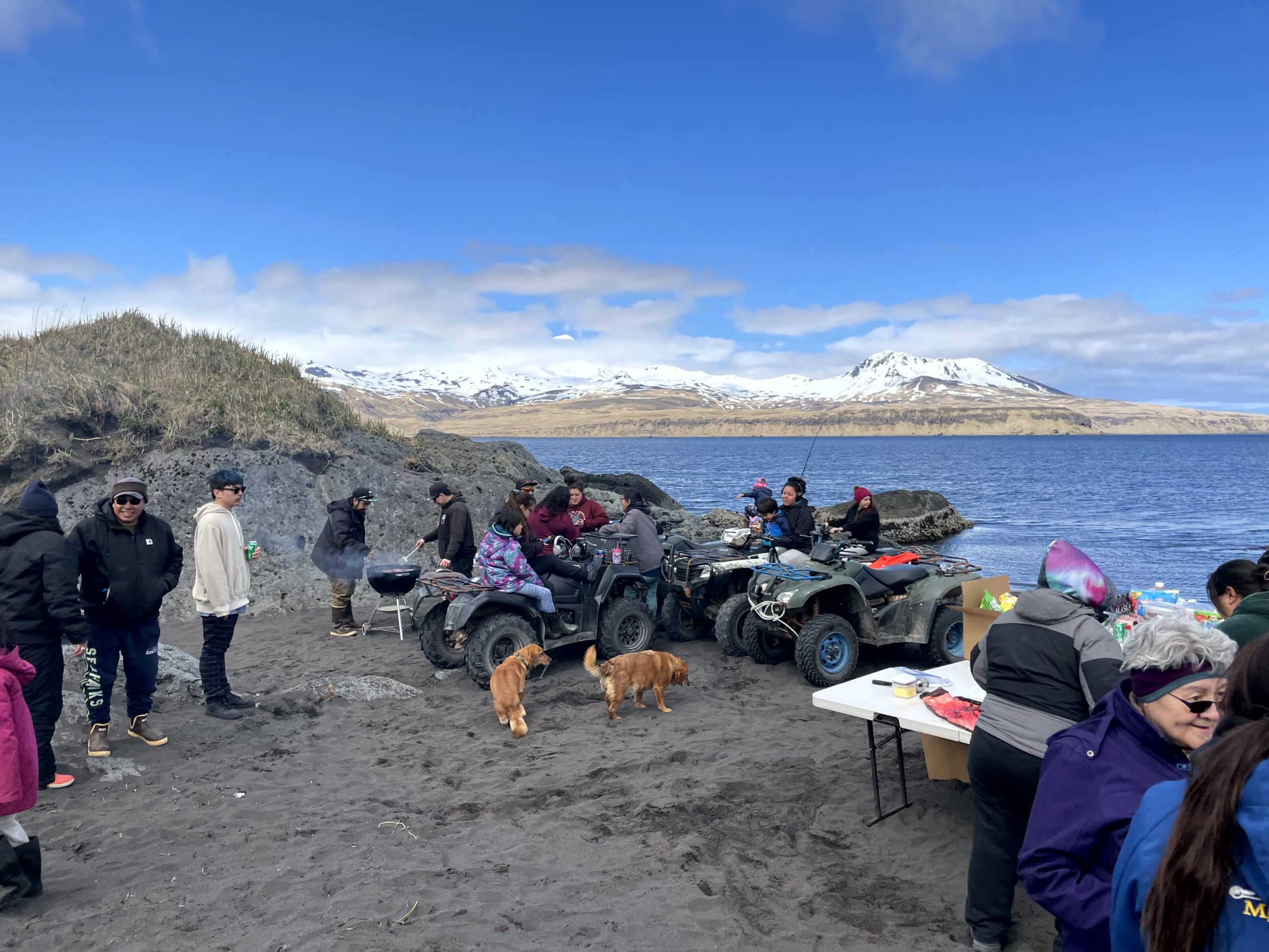 A group of people stand among ATV's and a grill. Behind them are snow-capped mountains and a calm, blue ocean. The sky is partially cloudy and very blue.