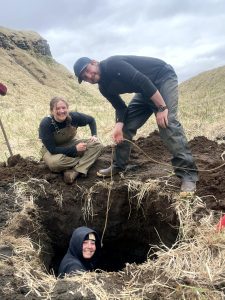 Two people stand and sit around the edge of a freshly dug hole, a third person is in the hole. Reyce is holding what looks like a thin rope. Behind them are grassy hills and an overcast sky.