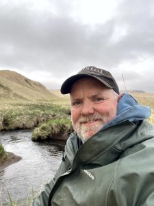 Chris taking a selfie with a creek, grassy hills, and an overcast sky as his background