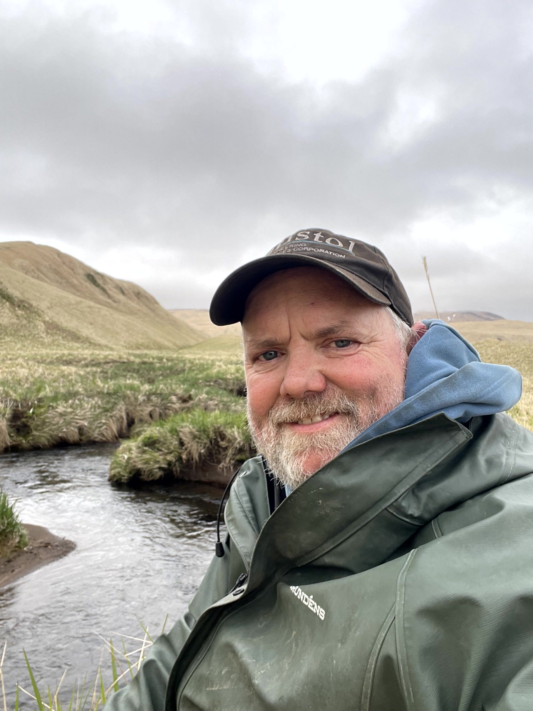 Chris taking a selfie with a creek, grassy hills, and an overcast sky as his background