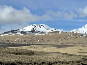 A landscape of rolling grassy foothills and snow-capped mountains. The sky is partially cloudy and blue.