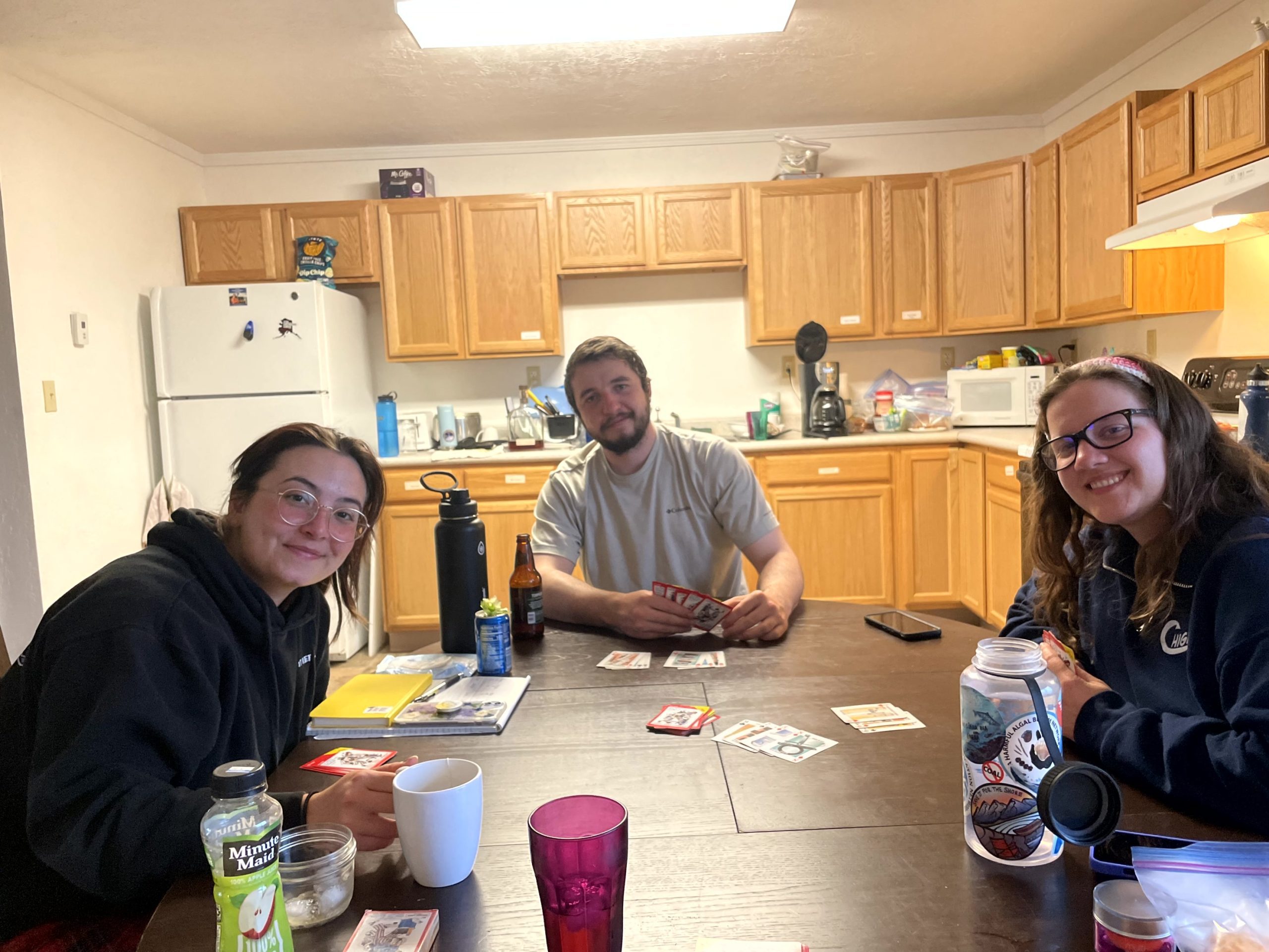 Three people sit at a table with playing cards. The background looks like someone's kitchen.