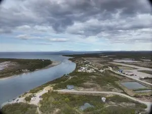 An aerial photo capturing part of Igiugig and a river running next to it. The camera is angled in a way that further out you can see the ocean. The sky is partially cloudy.