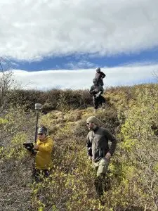 Four people stand in frame. One of them is holding the Trimble instrument. Two people are standing on the bottom of a slope in some brush. The other two are standing towards the top of the slope, one of them is holding a field notebook. The sky is partially cloudy.