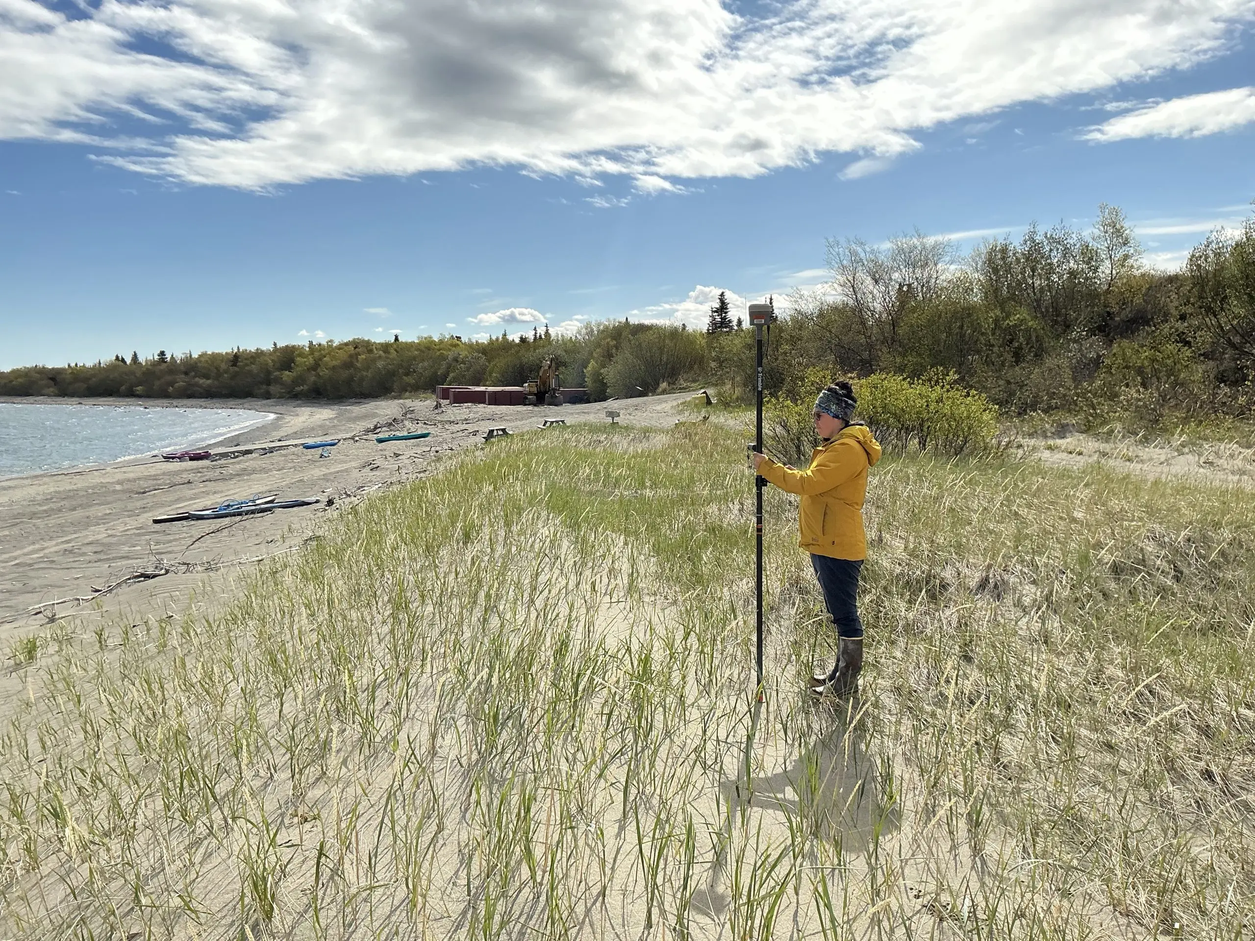 Someone stands on a grassy and sandy part of a beach looking towards the water on the left of the image. There is brush in the background. The sky is partially cloudy.