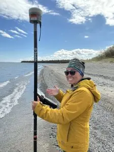 Someone stands holding a Trimble instrument. One hand is being held up with the palm facing the camera and the person is looking at the camera and smiling. They are standing on a beach near the water. There is brush in the background. The sky is mostly blue with a few clouds.