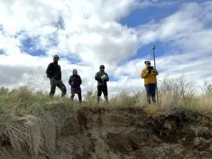 Four people stand on the top of a grassy ledge. One of them is holding a Trimble instrument, another is holding a field notebook. The sky is partially cloudy.