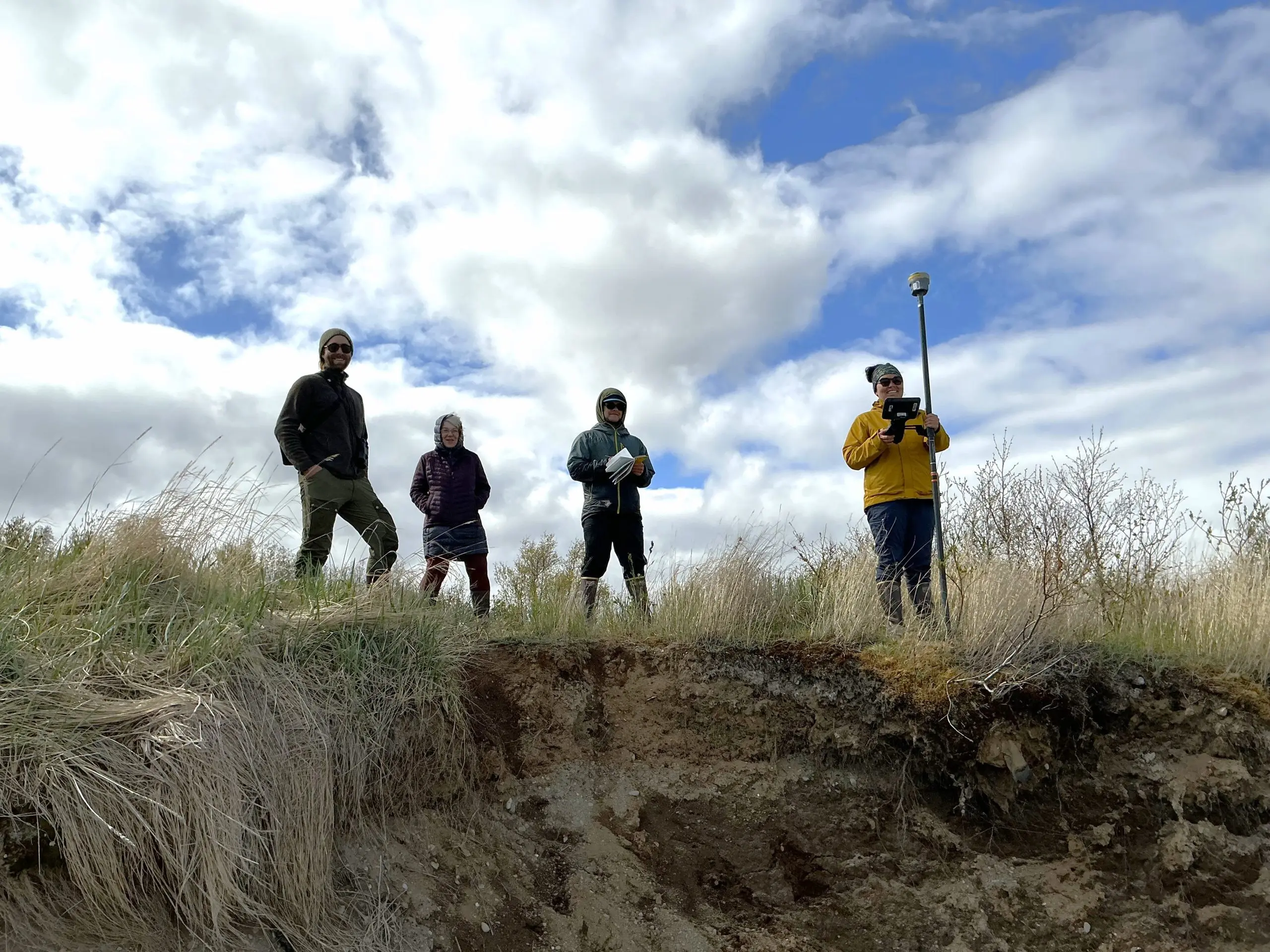 Four people stand on the top of a grassy ledge. One of them is holding a Trimble instrument, another is holding a field notebook. The sky is partially cloudy.