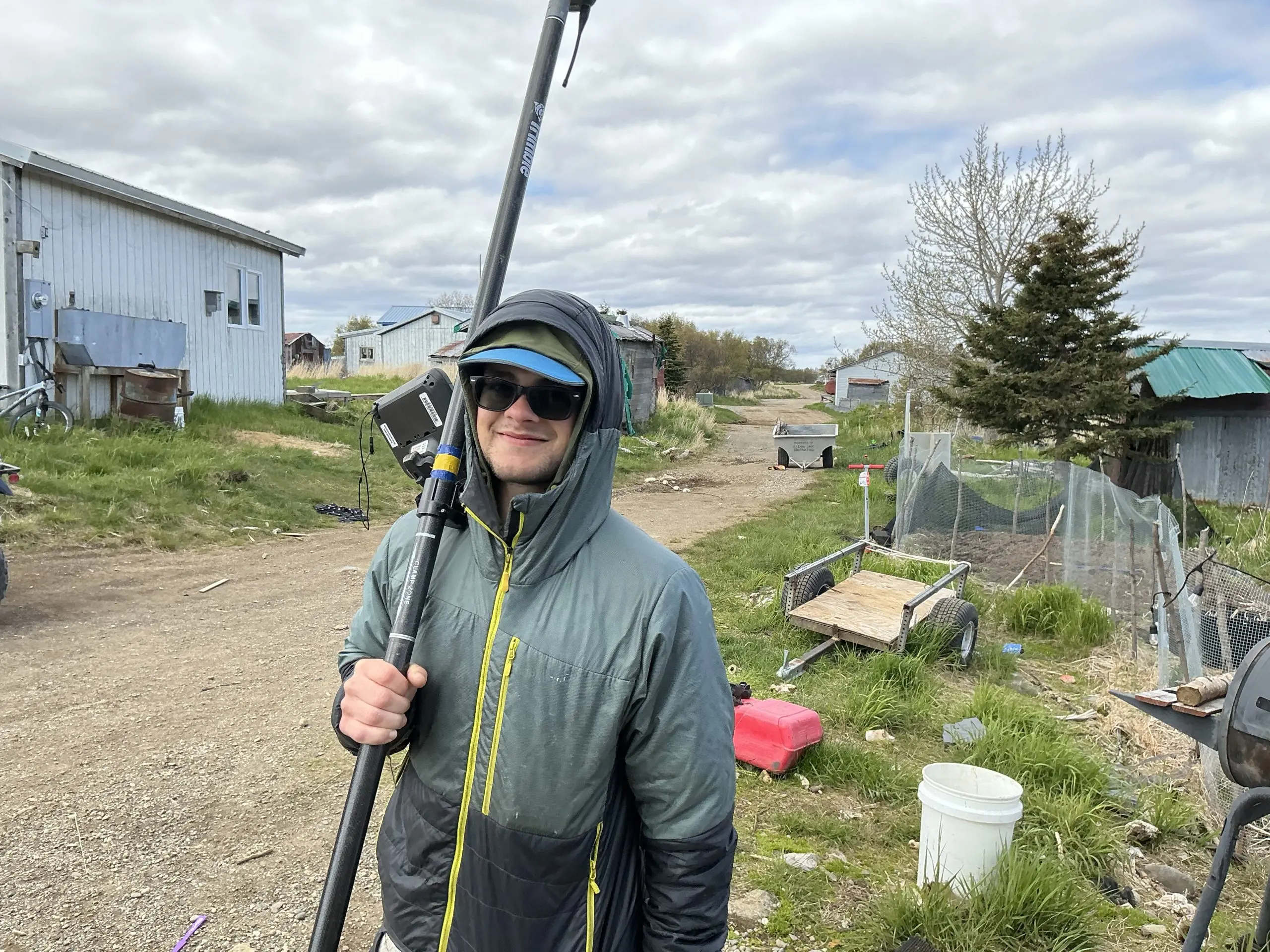Seth Classen is holding a Trimble instrument and looking towards the camera smiling. He is standing on the side of a dirt road in a neighborhood. The grass on either side of the road is green and various objects are scattered around. The sky is cloudy.