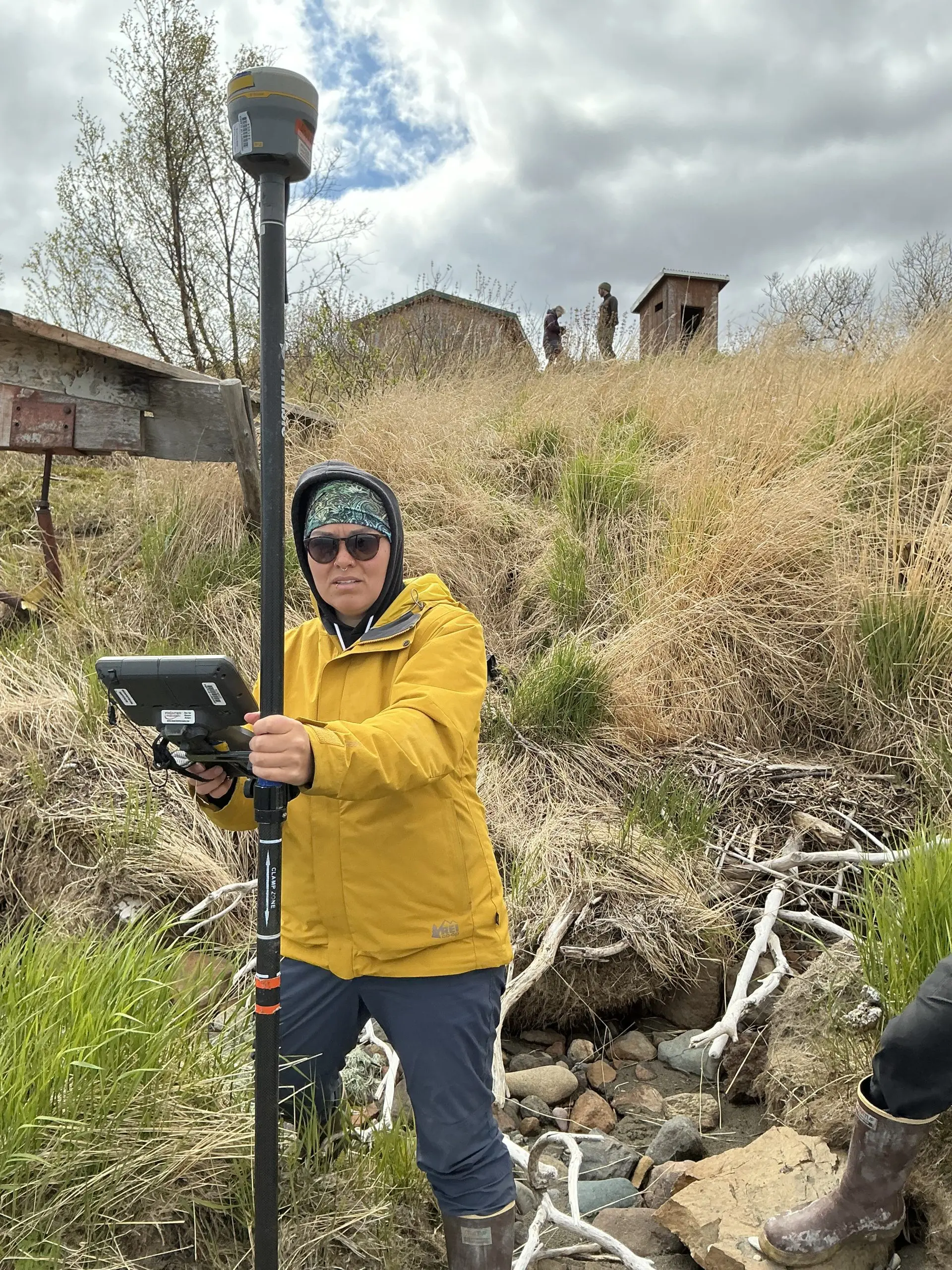 Someone stands in the middle of the photo holding a Trimble instrument. They are standing at the base of a grassy hill. There are a few buildings around the top of the hill. The sky is mostly cloudy.