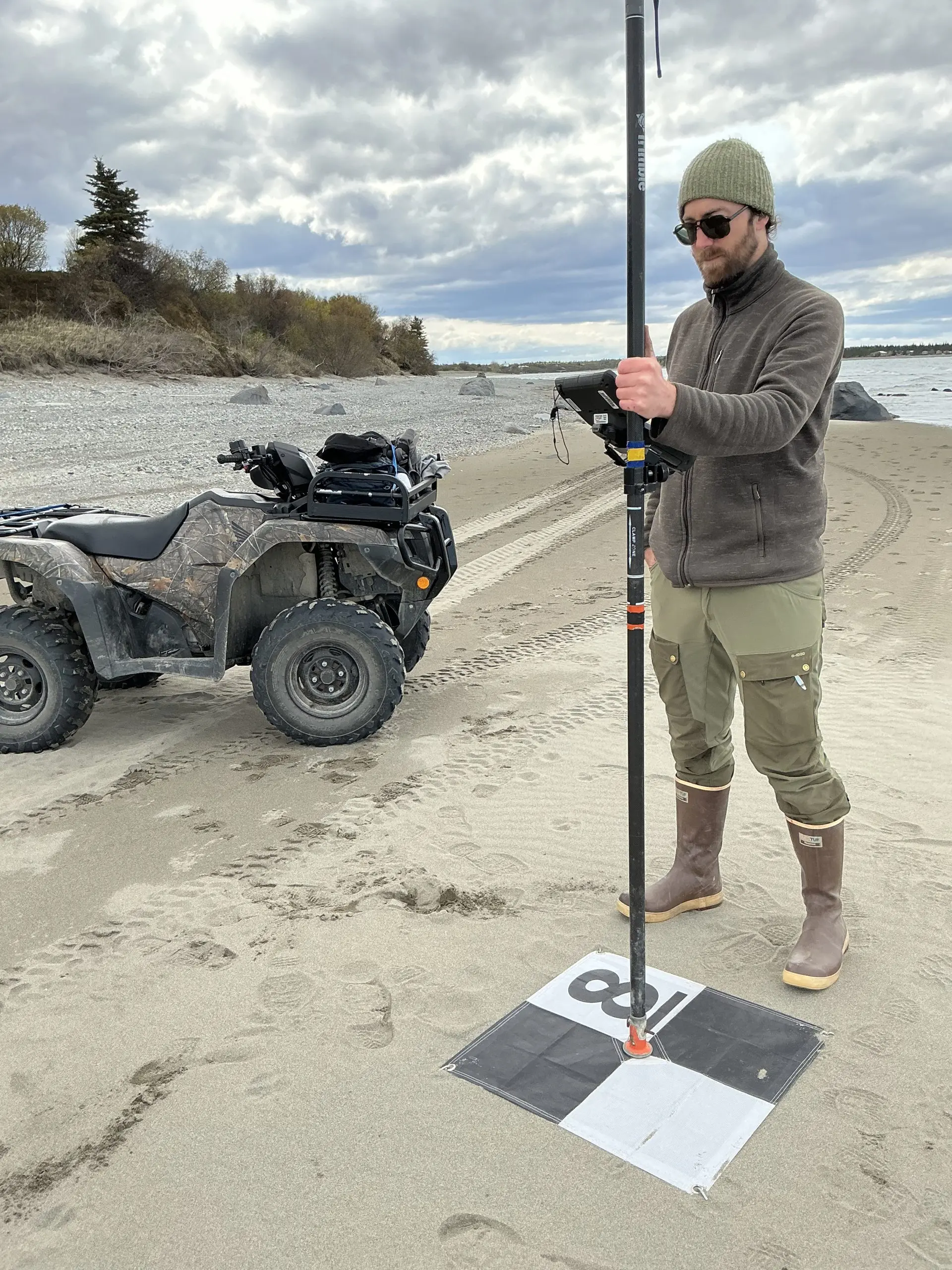 Someone stands holding a Trimble instrument on a GCP tarp. To the left of the photo is a parked ATV. They are on the sandy area of a beach. Further back there is some gravel and a brushy hill behind the gravel. The sky is cloudy.