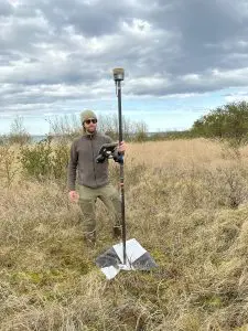 Someone stands in a grassy field holding a Trimble instrument on a GCP tarp. The sky is mostly cloudy.