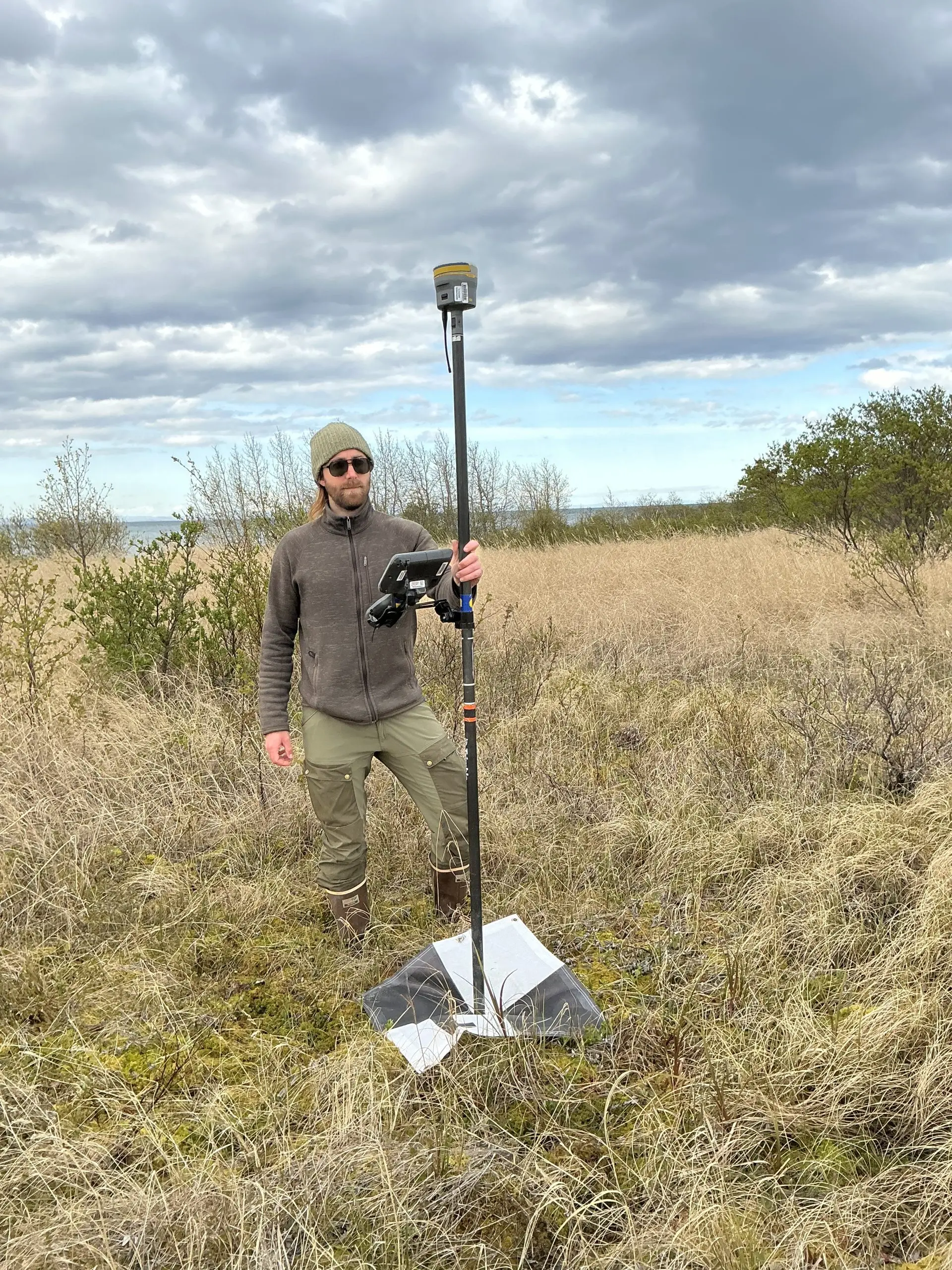 Someone stands in a grassy field holding a Trimble instrument on a GCP tarp. The sky is mostly cloudy.