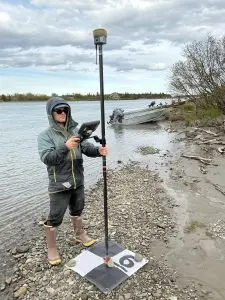 Seth Classen is holding a Trimble instrument on a GCP tarp. He is standing on a gravely section of a beach. The water is calm. There is a metal boat parked behind him, leaning on a grassy shore. There are trees and a house on the far side of the water. The sky is cloudy.
