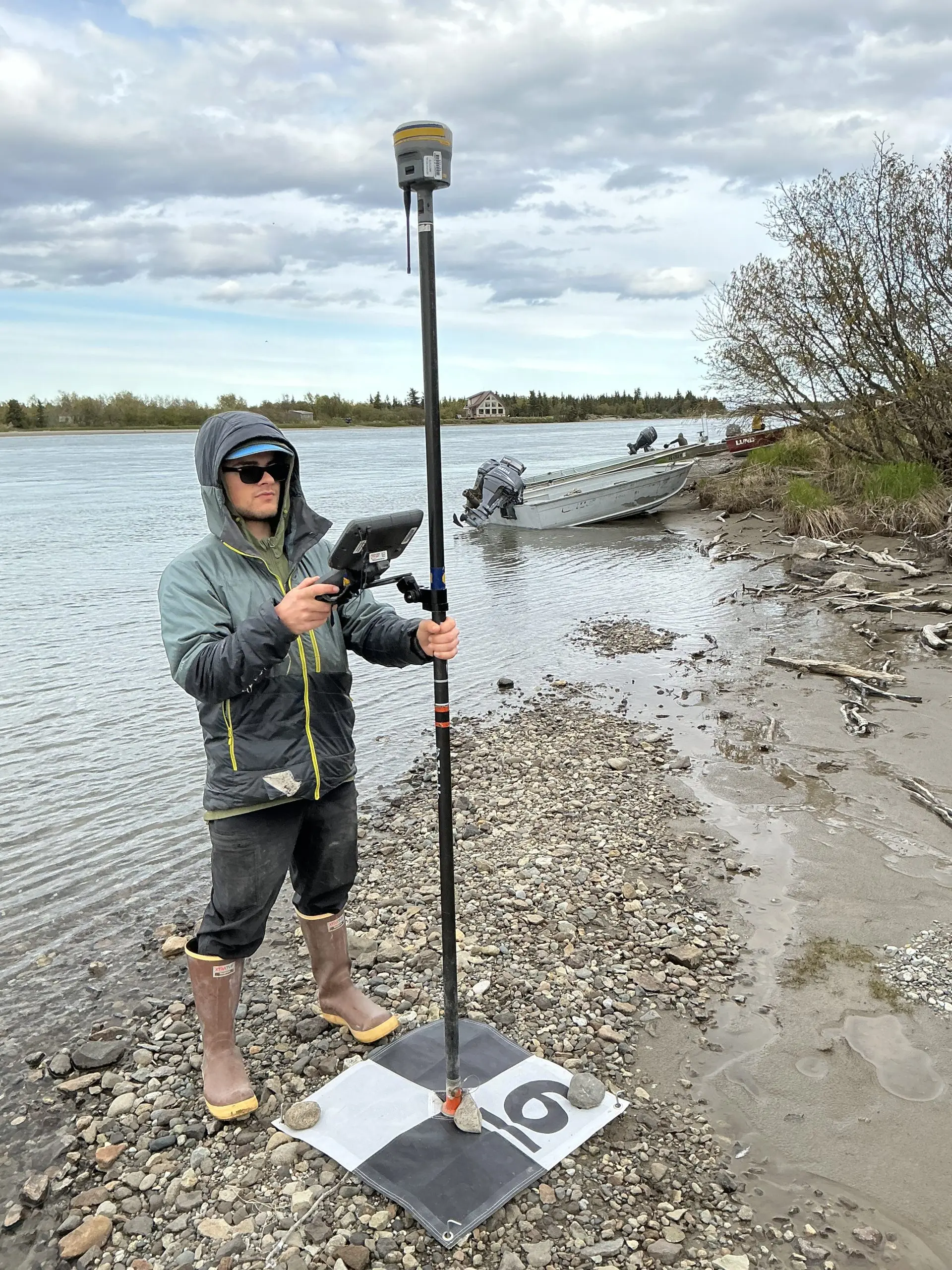 Seth Classen is holding a Trimble instrument on a GCP tarp. He is standing on a gravely section of a beach. The water is calm. There is a metal boat parked behind him, leaning on a grassy shore. There are trees and a house on the far side of the water. The sky is cloudy.