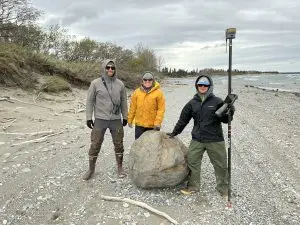 Three people stand around a boulder on the beach. One of them is holding a Trimble instrument. Two of them are touching the boulder. The beach is gravelly with bits of driftwood scattered about. There is a small bluff to the left of the frame covered in brush. There are small waves forming in the water to the right of the frame. The sky is overcast.