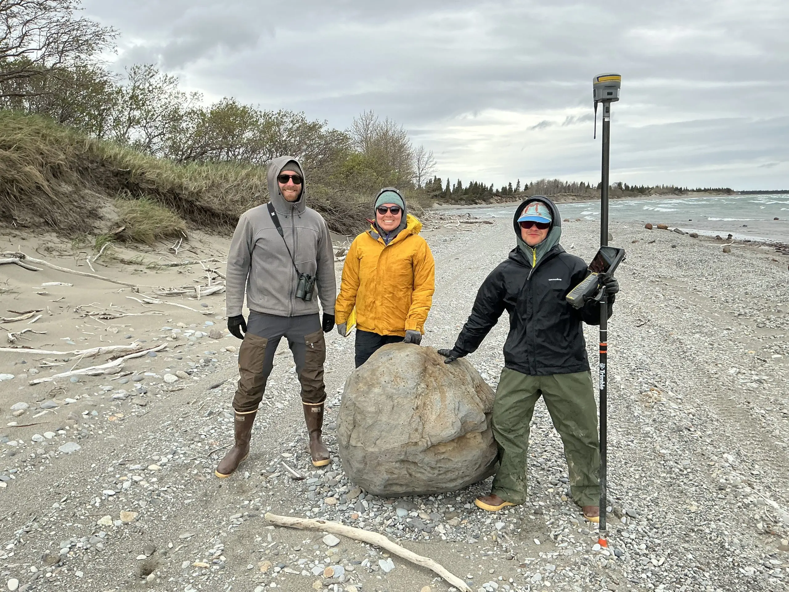Three people stand around a boulder on the beach. One of them is holding a Trimble instrument. Two of them are touching the boulder. The beach is gravelly with bits of driftwood scattered about. There is a small bluff to the left of the frame covered in brush. There are small waves forming in the water to the right of the frame. The sky is overcast.
