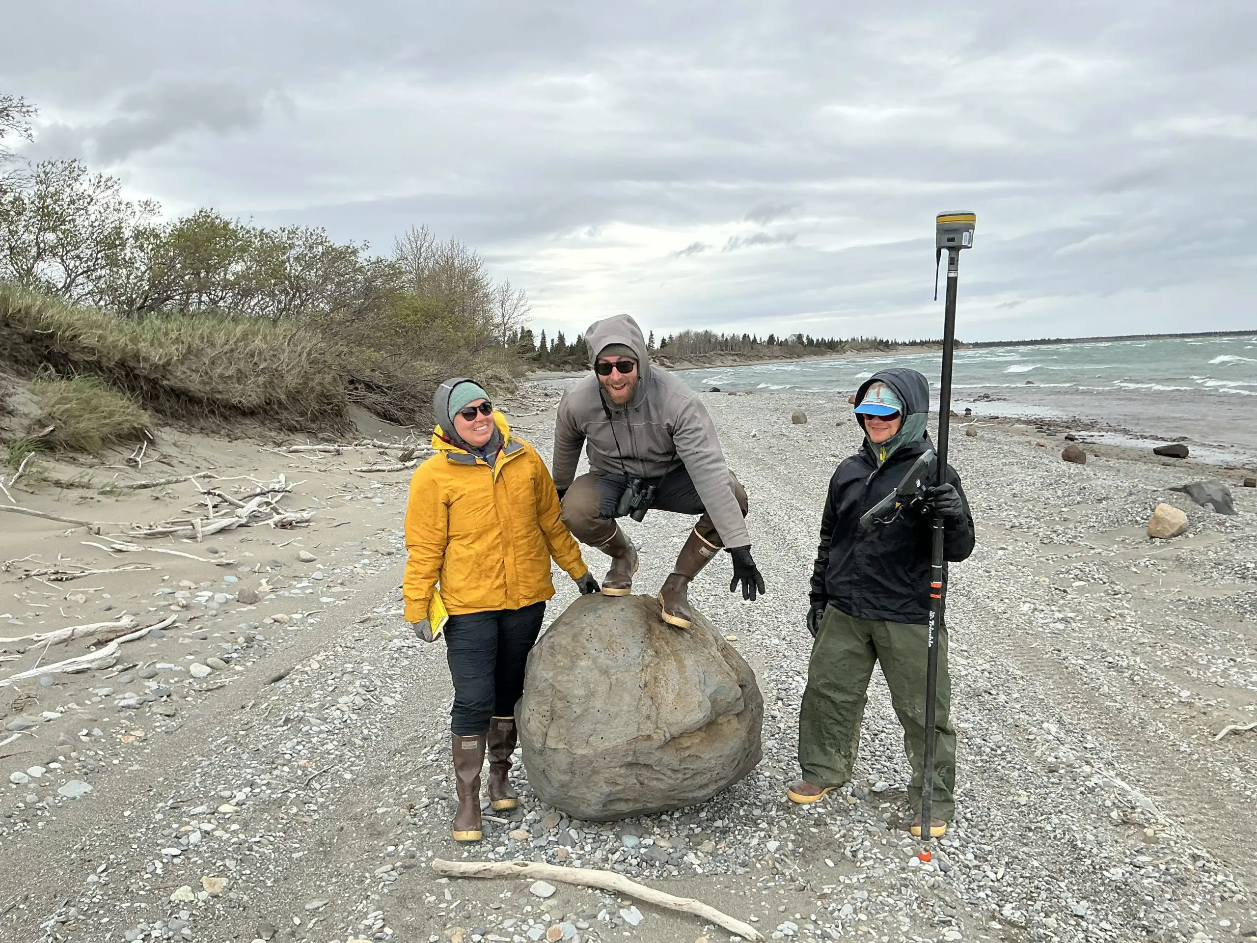 Three people are on a beach with a boulder. One of them is holding a field notebook with their hand on the boulder, another is squat-standing on the boulder, and the other is standing on the other side of the boulder holding a Trimble instrument. The beach is gravelly with some driftwood scattered around. The left has a small bluff that's covered in brush. The right is the ocean with small waves breaking. The sky is overcast.