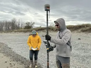 Two people are standing on a gravelly beach. One of them is holding a Trimble instrument and the other is looking into their open field notebook. Behind them is a small bluff covered in tall grass. There are leafless trees in the background to the left of the frame. The sky is overcast.