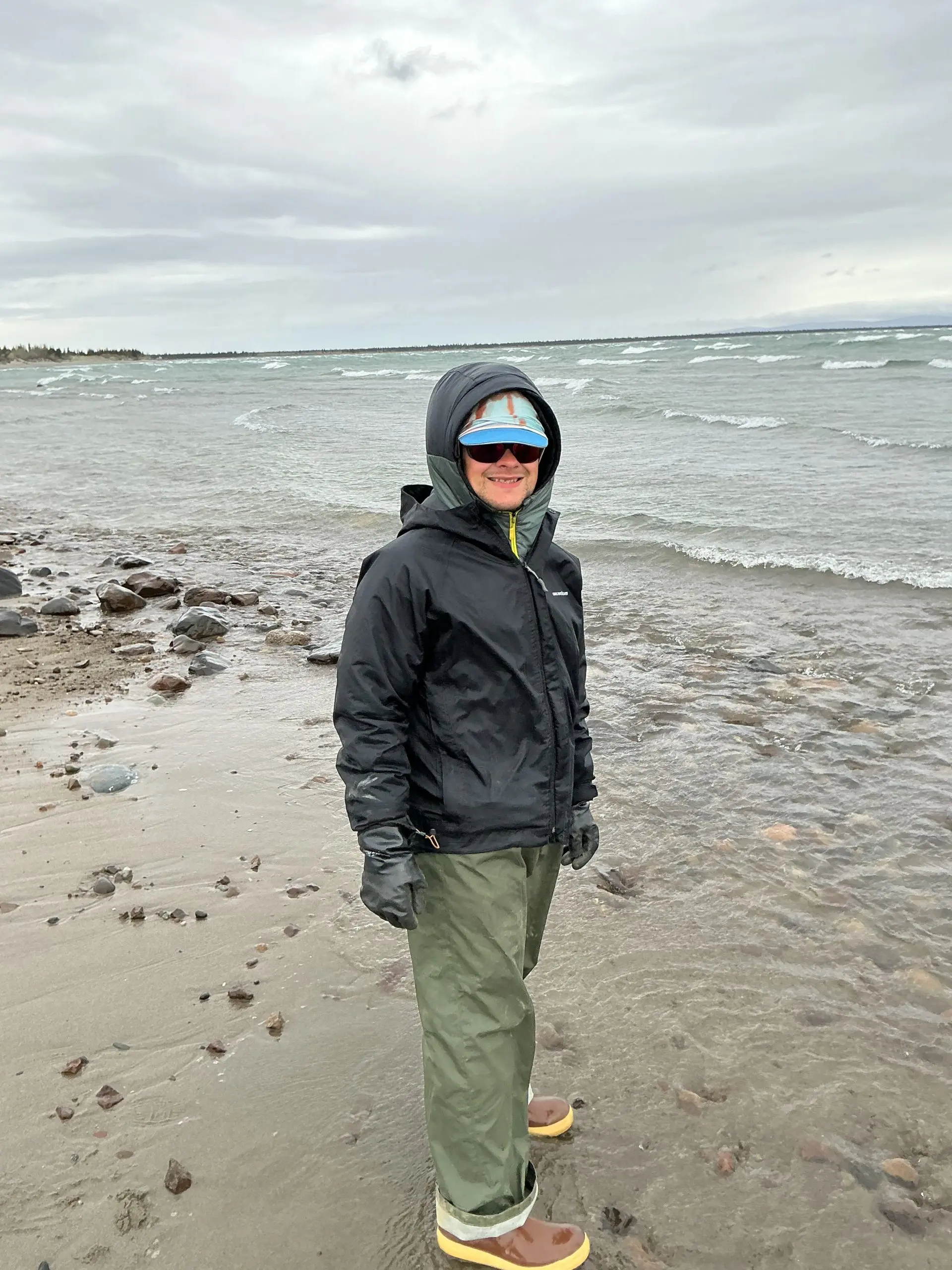 Seth Classen is standing on a beach with beach water receding around his feet. There are a few rocks scattered around the sand. The deeper water has small waves breaking. The sky is overcast.