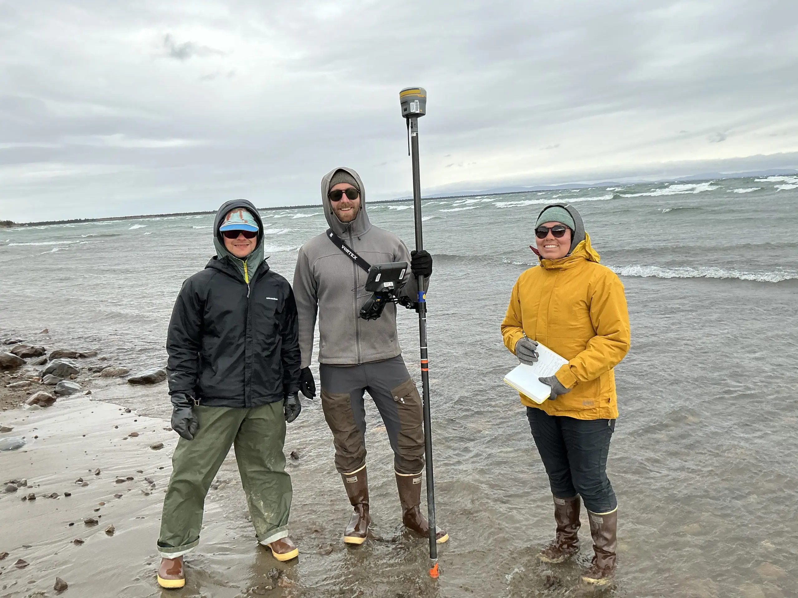 Three people stand on a beach with ocean water receding around their feet. One of them is holding a Trimble instrument, another is holding an open notebook. There area few rocks scattered around the sand. The deeper water has small waves breaking. The sky is overcast.