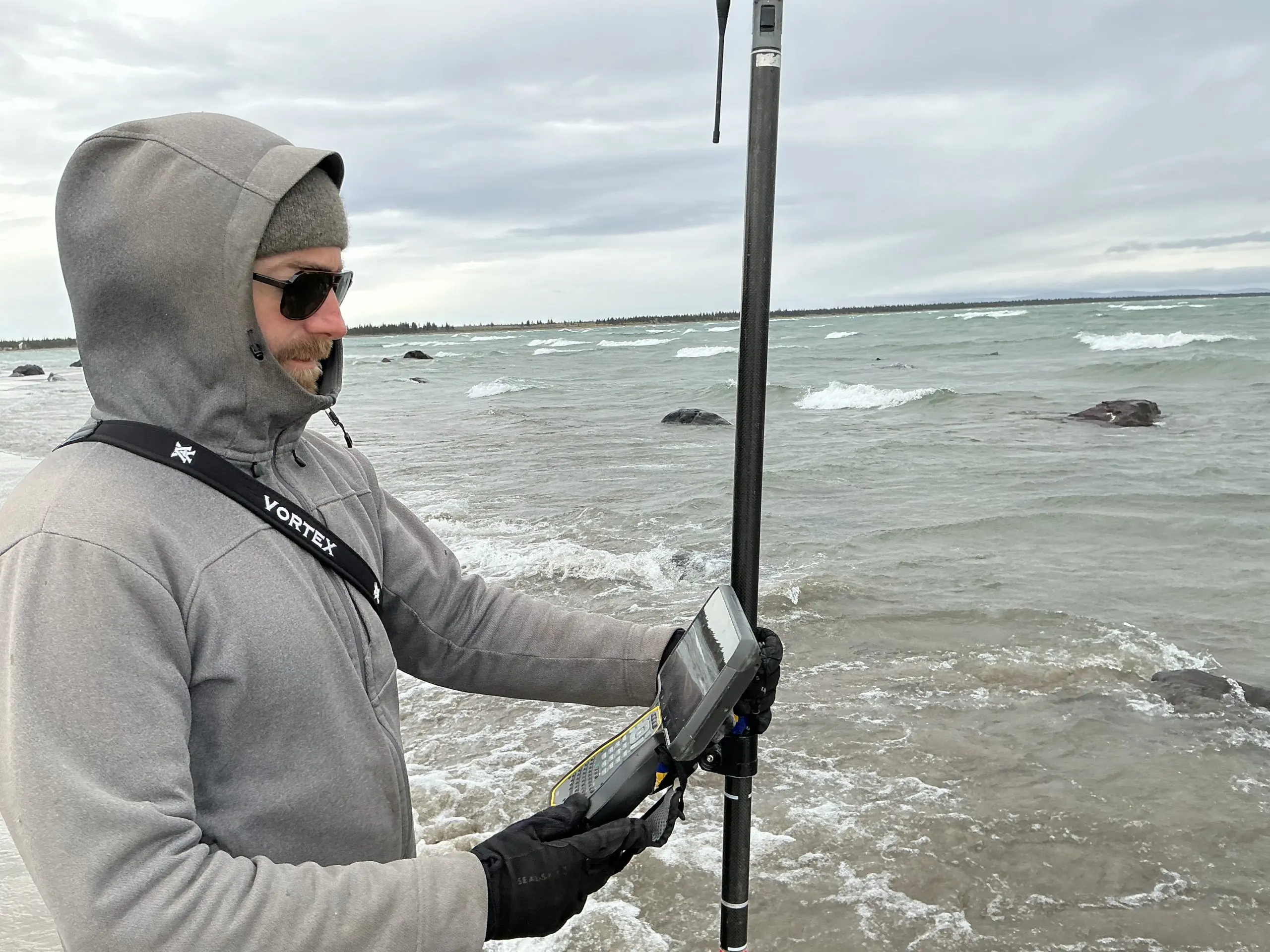 Someone is holding a Trimble instrument, looking at the small screen on it. They seem to be in shallow beach water. The background is choppy seawater. The sky is overcast.