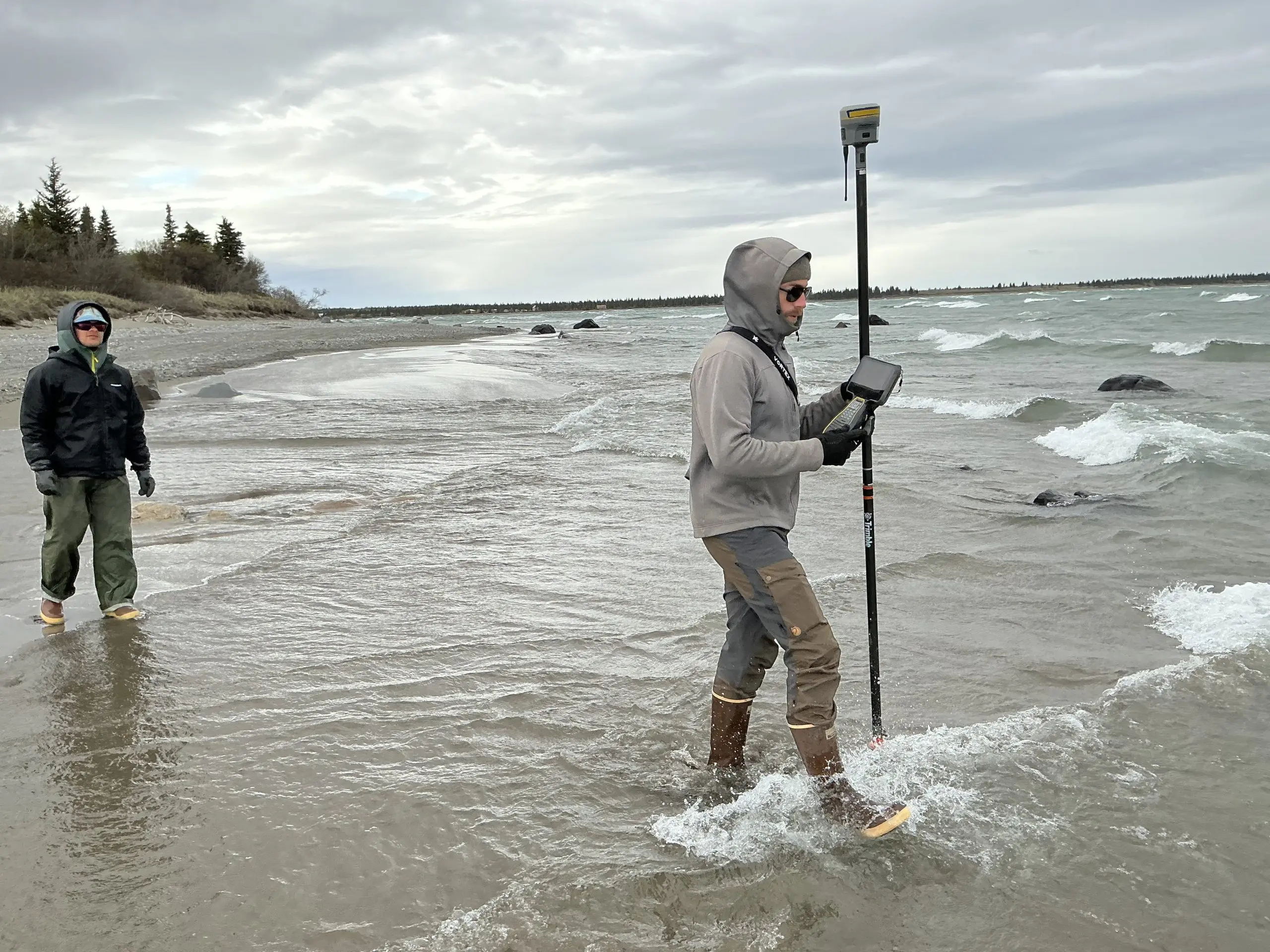 Two people are standing in shallow water on a beach. One of them is holding a Trimble instrument and water is splashing by their feet. The other is in the left of the frame where the water starts to recede. Further back there is a beach with some driftwood, brush, and trees. The sky is overcast.