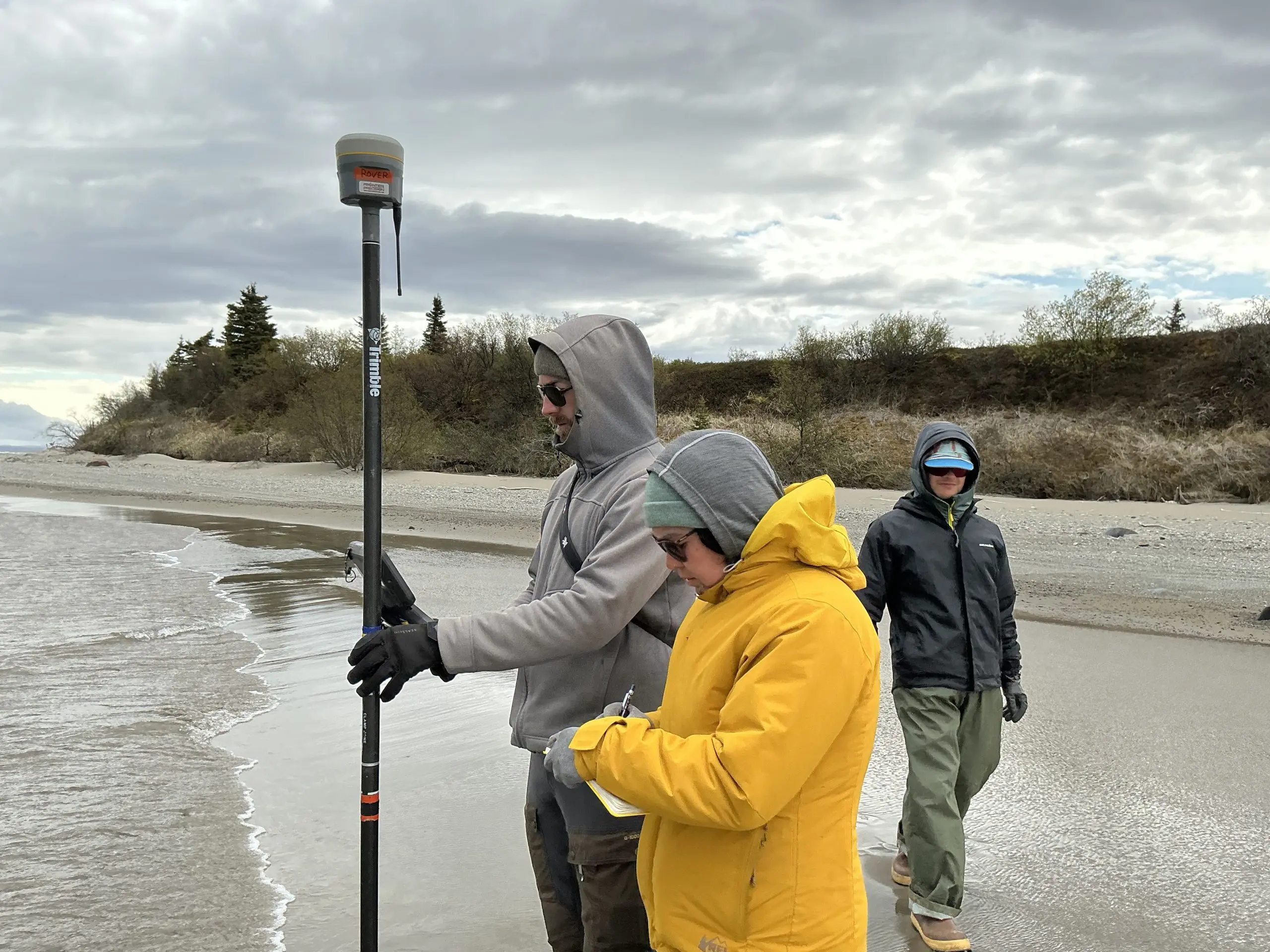 Three people stand on a beach. One of them is holding a Trimble instrument and another is writing in a field notebook. The two with the Trimble and the notebook are standing on the edge of the water. Behind them is a small hill with thick brush. The sky is cloudy.