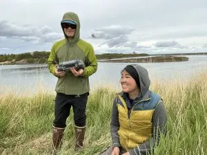 Two people are in frame. One of them is holding drone controllers while the other is kneeling on the ground. They are in tall grass. Behind them is a calm body of water. The other side of the water has low hills and trees. The sky is overcast.