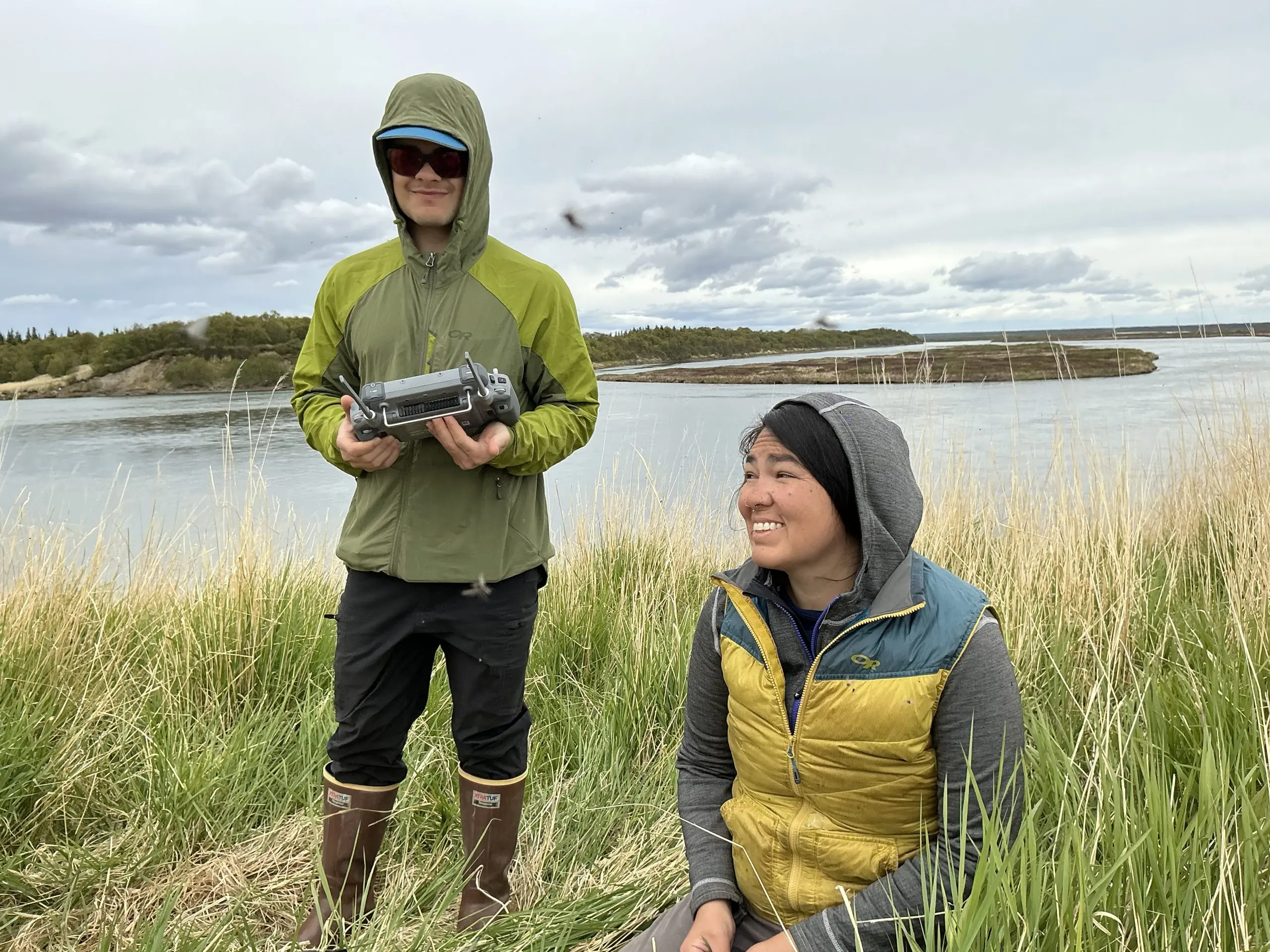 Two people are in frame. One of them is holding drone controllers while the other is kneeling on the ground. They are in tall grass. Behind them is a calm body of water. The other side of the water has low hills and trees. The sky is overcast.