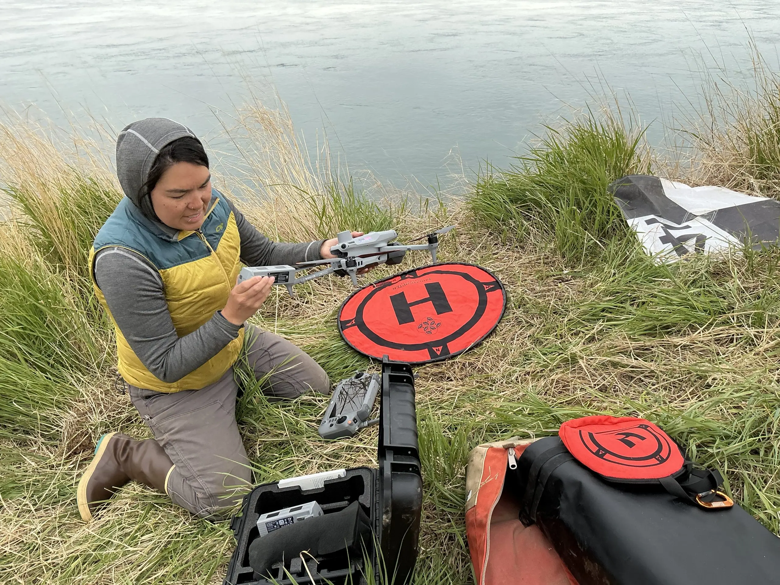 Someone knees in tall grass holding a drone. In front of them are drone controllers, a landing pad, a GCP tarp, and a few bags. Behind them is calm water with some wind ripples.