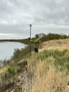 Seth Classen is standing on the downward slope of a grassy hill holding a Trimble instrument. The background has taller brush and calm water. The sky is overcast.