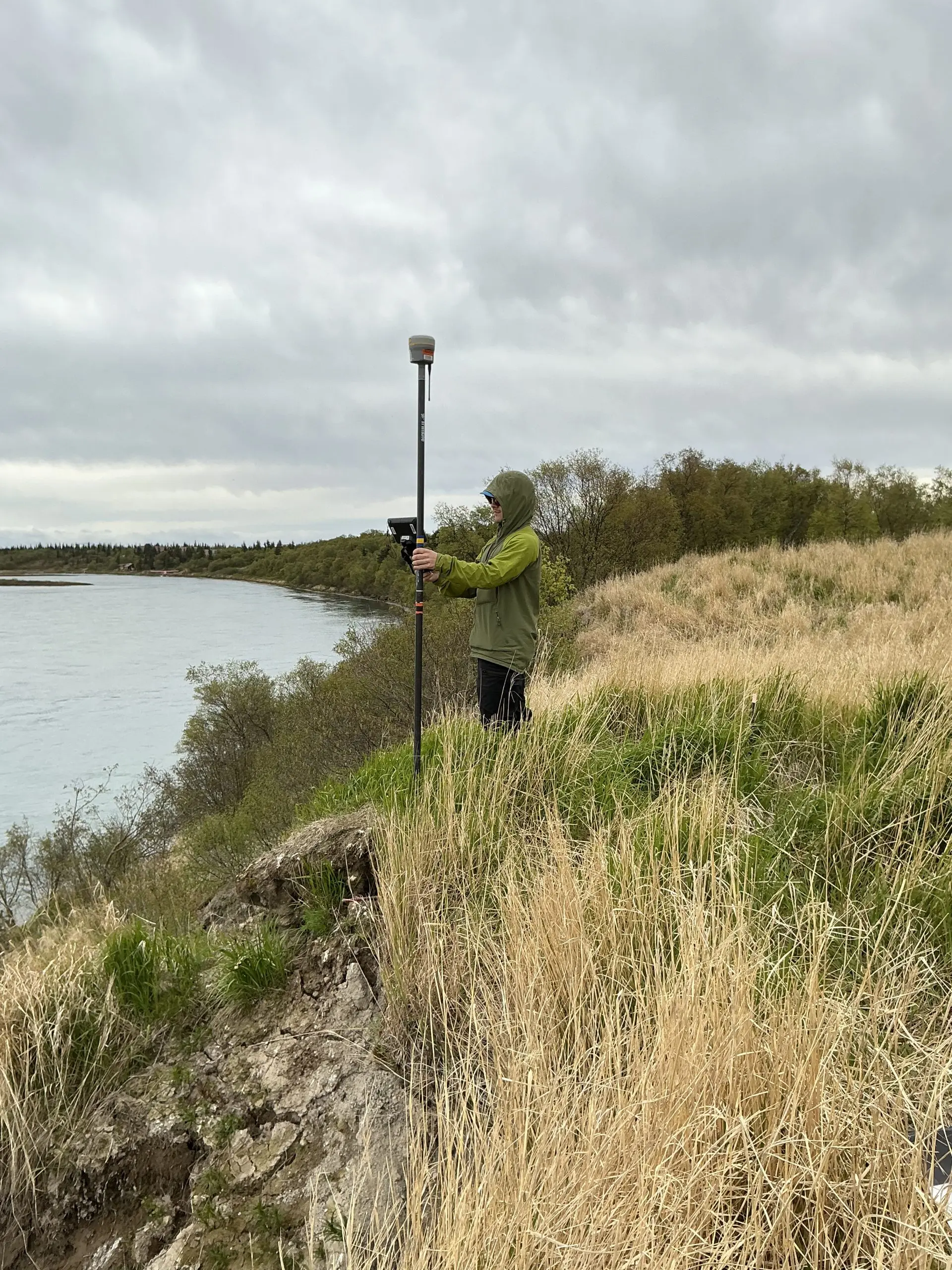 Seth Classen is standing on the downward slope of a grassy hill holding a Trimble instrument. The background has taller brush and calm water. The sky is overcast.