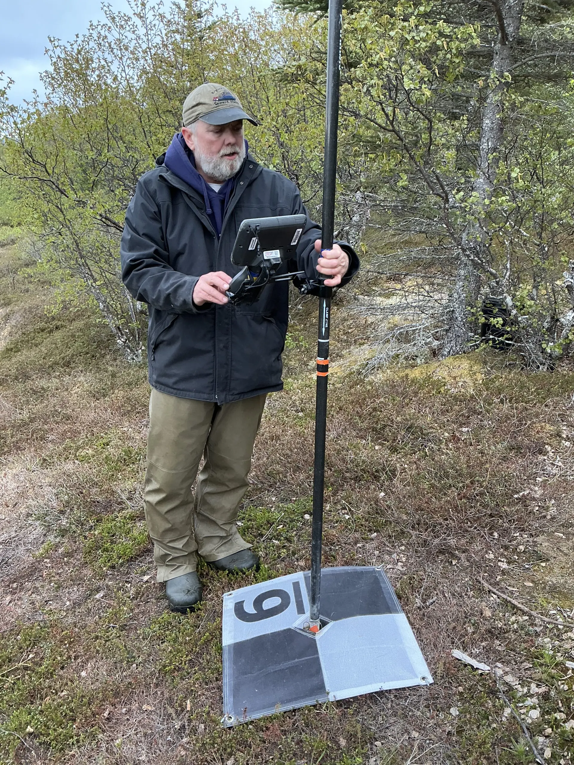 Chris Maio is holding a Trimble instrument on a GCP tarp. They are standing on a small clearing on the edge of a brush line. The sky is partially cloudy.