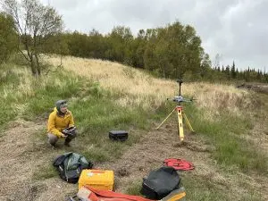Someone squats while holding controllers for a drone. Near them are some bags, containers, a drone tarp, and a tripod. They are all on top of tall grass on a gently sloping hill. There are some trees further back. The sky is overcast. A drone is in flight, starting its ascent.