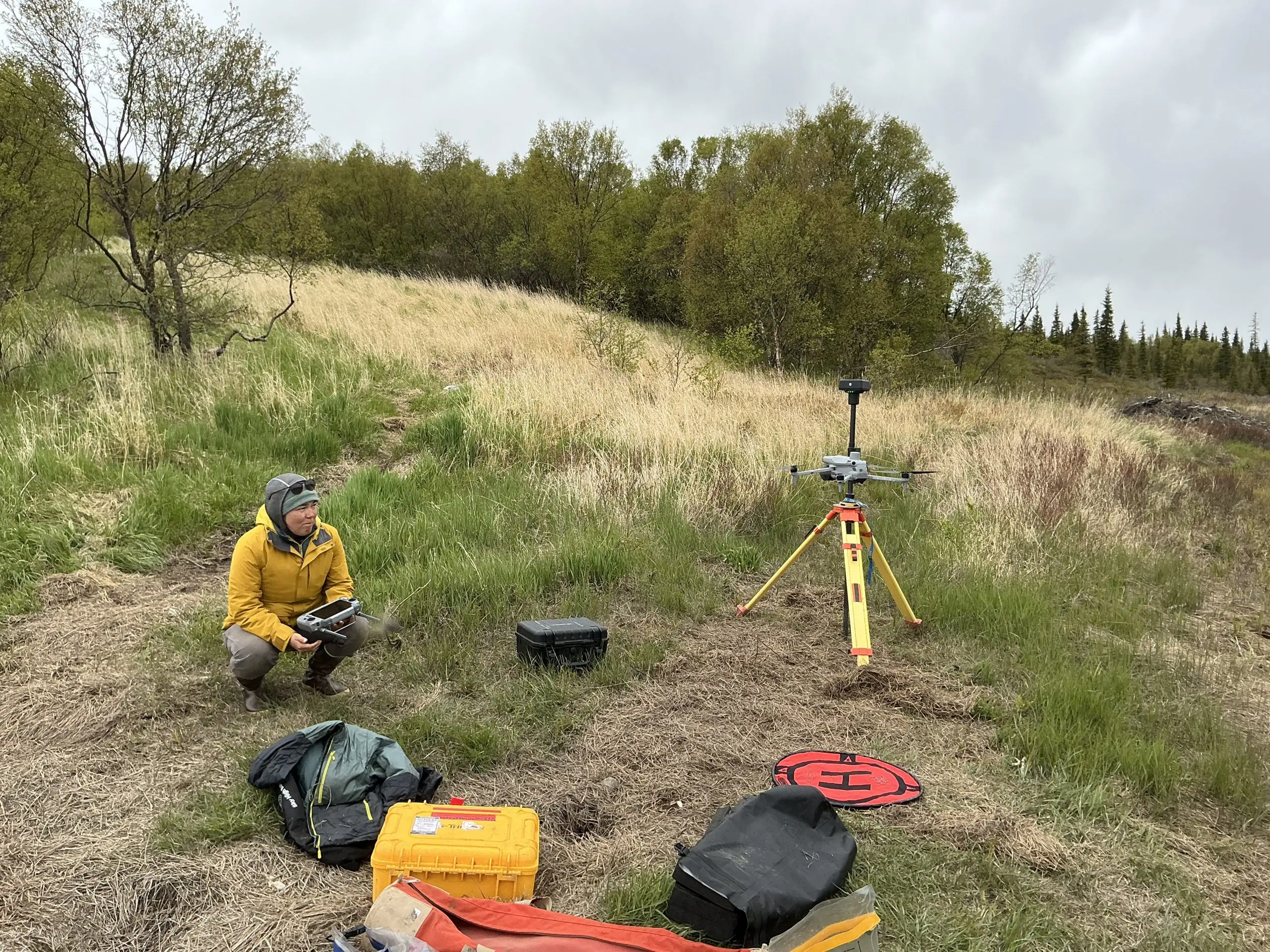 Someone squats while holding controllers for a drone. Near them are some bags, containers, a drone tarp, and a tripod. They are all on top of tall grass on a gently sloping hill. There are some trees further back. The sky is overcast. A drone is in flight, starting its ascent.