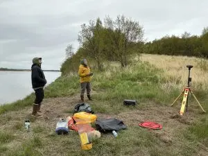 Two people stand near each other. One of them is holding controllers for a drone. They are both looking up towards something out of camera frame. There are some bags, containers, a drone landing tarp, and a tripod nearby. They are on a grassy hill near a cliff edge. There is smooth water visible off the side of the cliff. The sky is overcast.