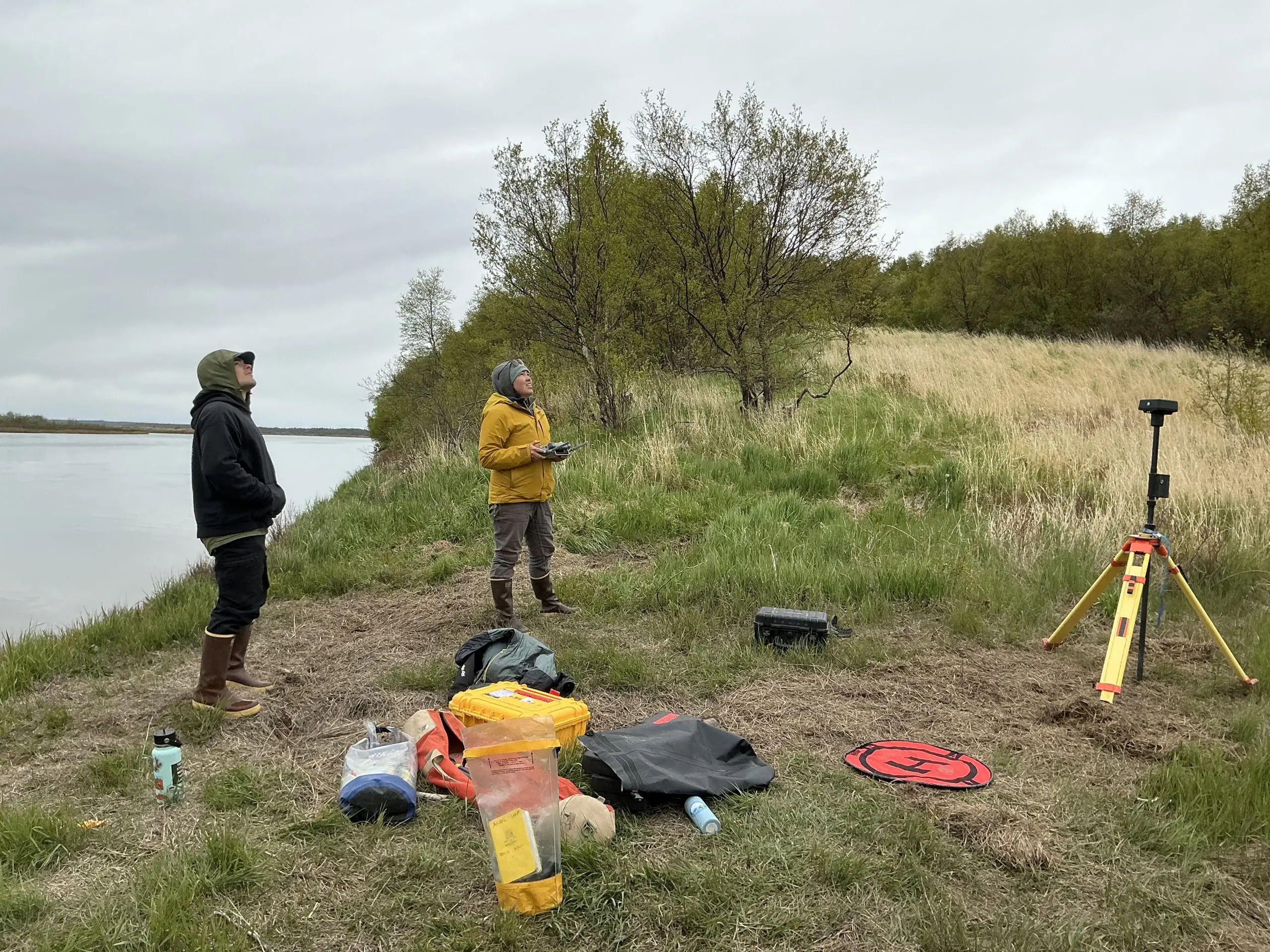 Two people stand near each other. One of them is holding controllers for a drone. They are both looking up towards something out of camera frame. There are some bags, containers, a drone landing tarp, and a tripod nearby. They are on a grassy hill near a cliff edge. There is smooth water visible off the side of the cliff. The sky is overcast.