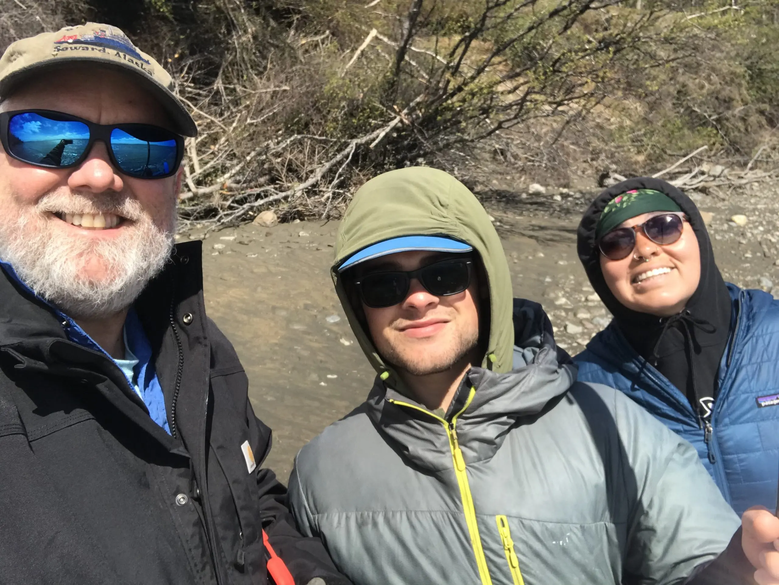 Three people stand near each other for a group selfie. Behind them there are some rocks, mud, and some brush.