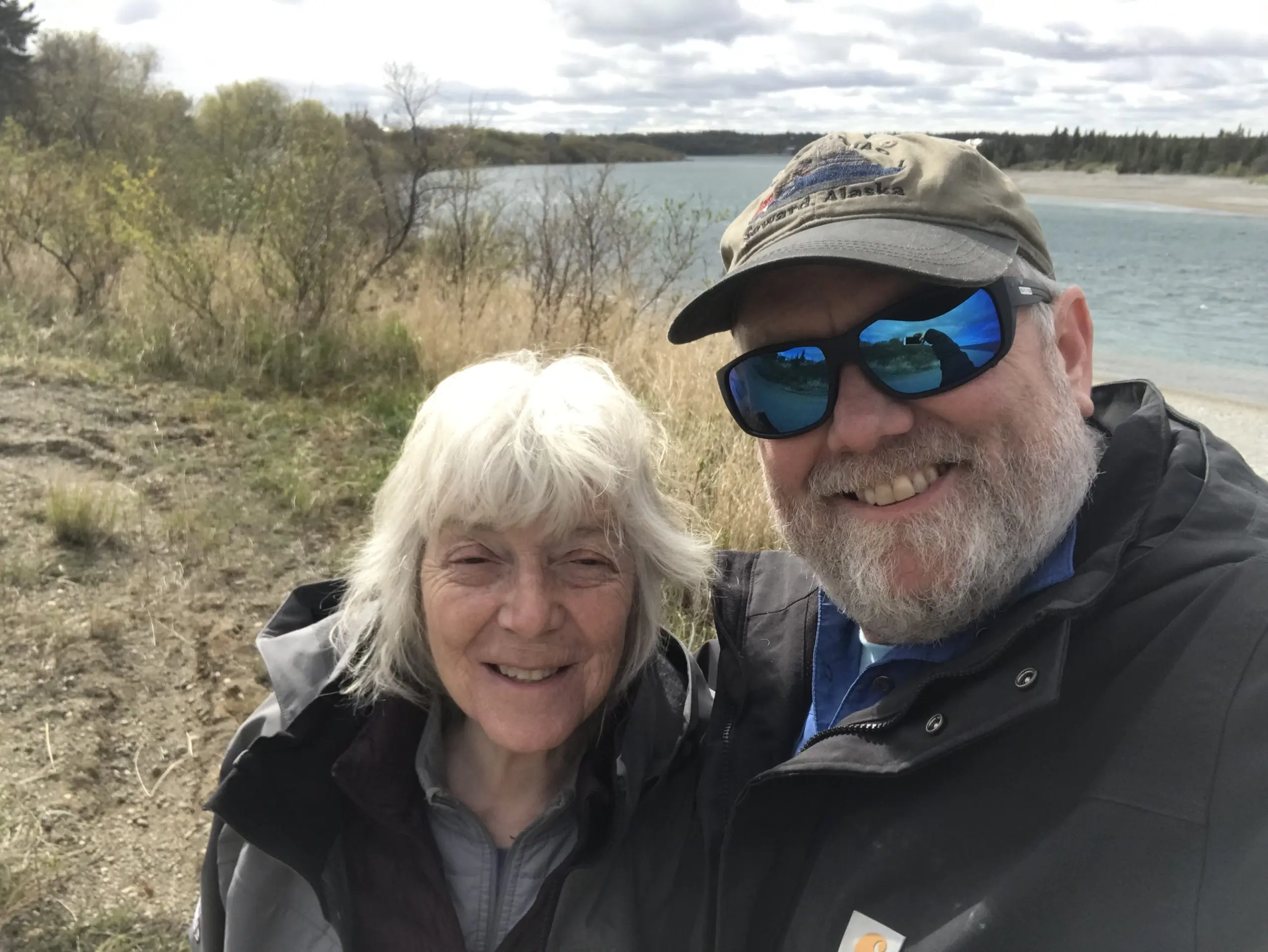 Two people stand next to each other for a selfie. Behind them there is a hill with brush and tall grass. Further back there is a river with evergreen trees on the opposite river bank.