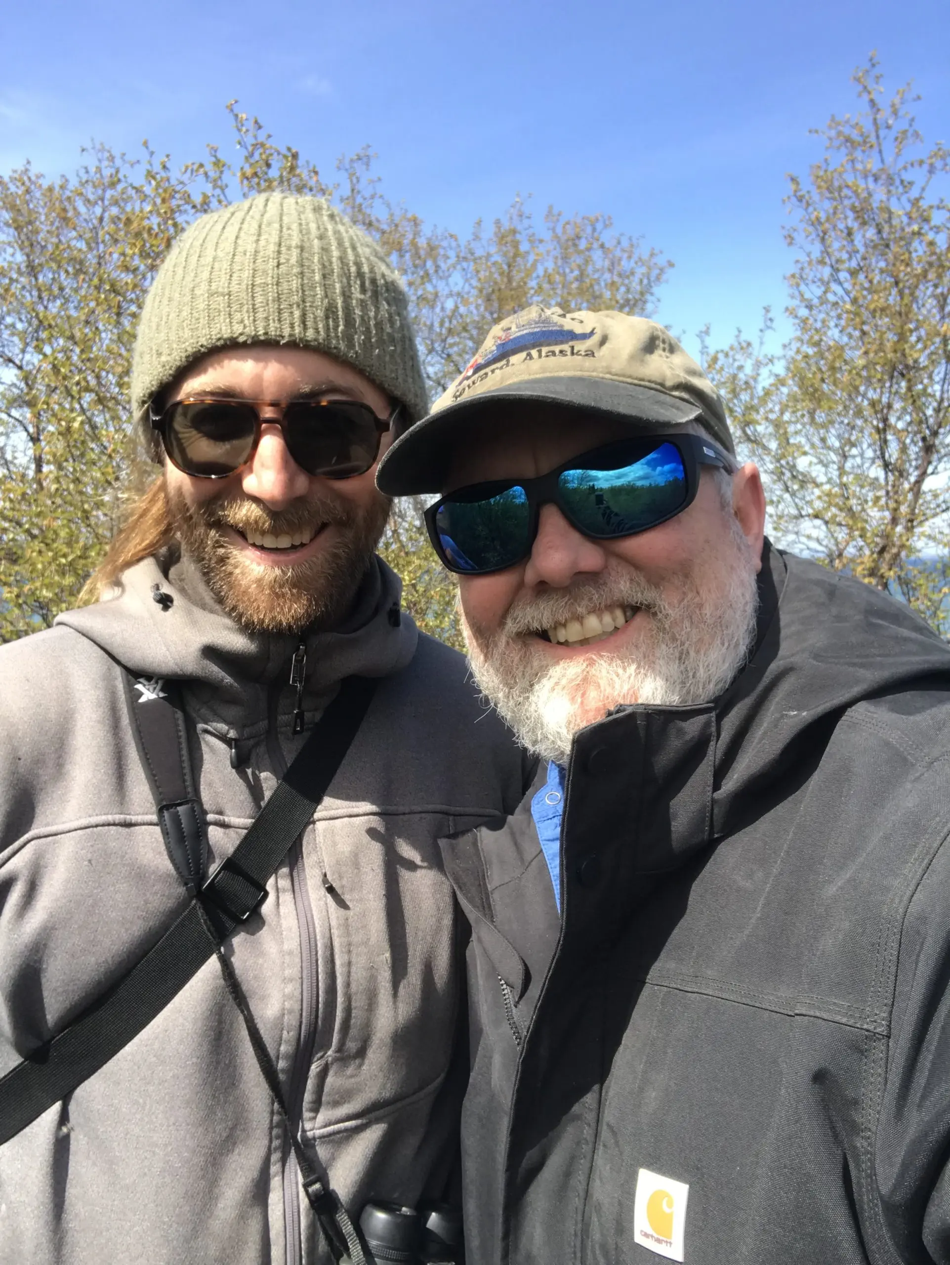 Two people stand next to each other for a selfie. There are some trees behind them and a blue sky.