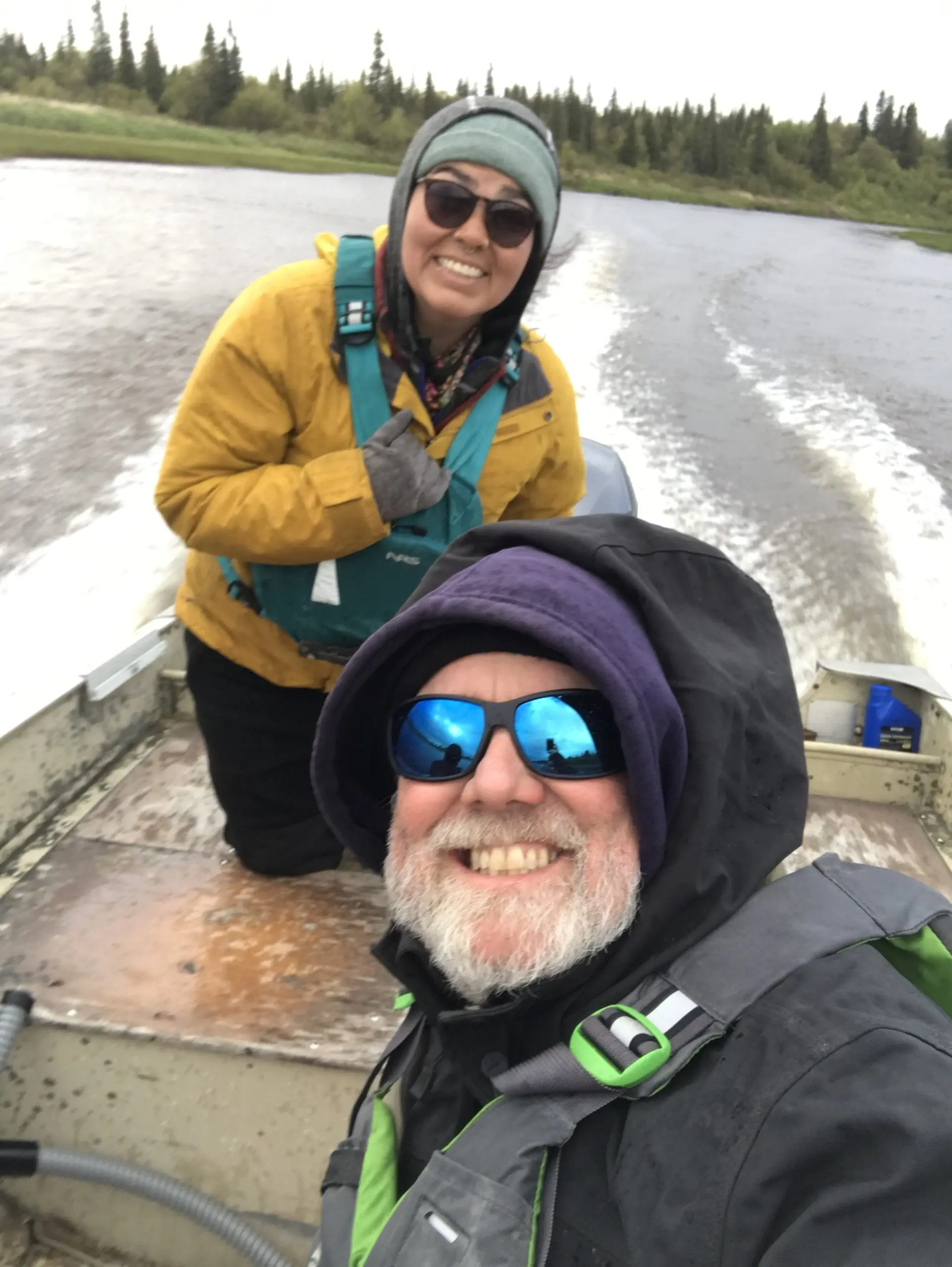 Two people sit in a moving boat looking toward the camera for a selfie. The water behind them is smooth with wakes forming. The river's shore is green with grass, brush, and evergreen trees.