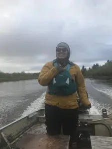 Someone stands near the back of the boat holding on to the rudder controller. Behind the boat are wakes on smooth water. The shores of the river are green with grass, brush, and trees.