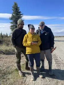 Three people stand near each other on the side of a graveled road. Two of them are looking at the phone screen being held by the third person. Behind them are a few evergreen trees and a partially cloudy sky.