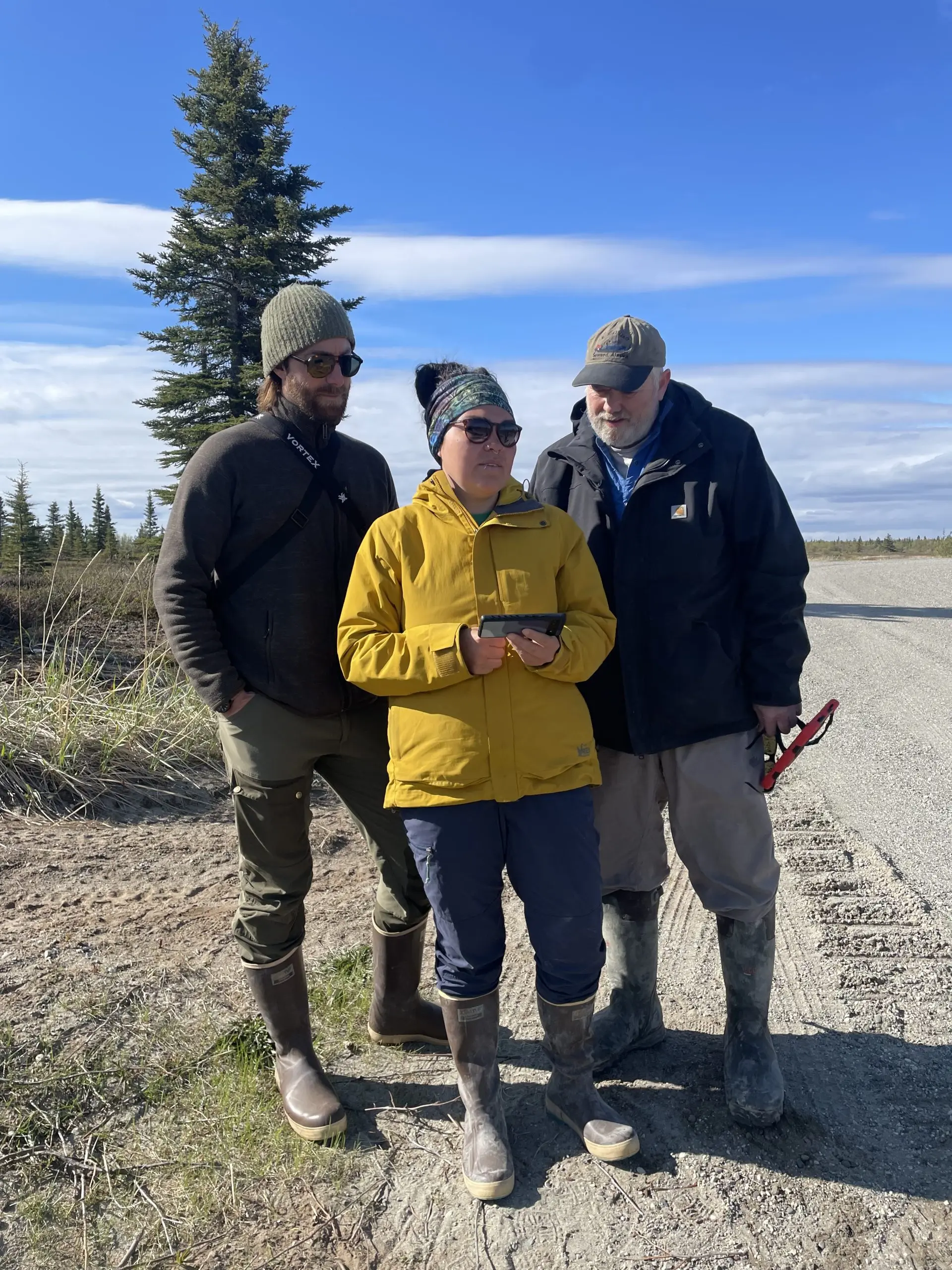 Three people stand near each other on the side of a graveled road. Two of them are looking at the phone screen being held by the third person. Behind them are a few evergreen trees and a partially cloudy sky.